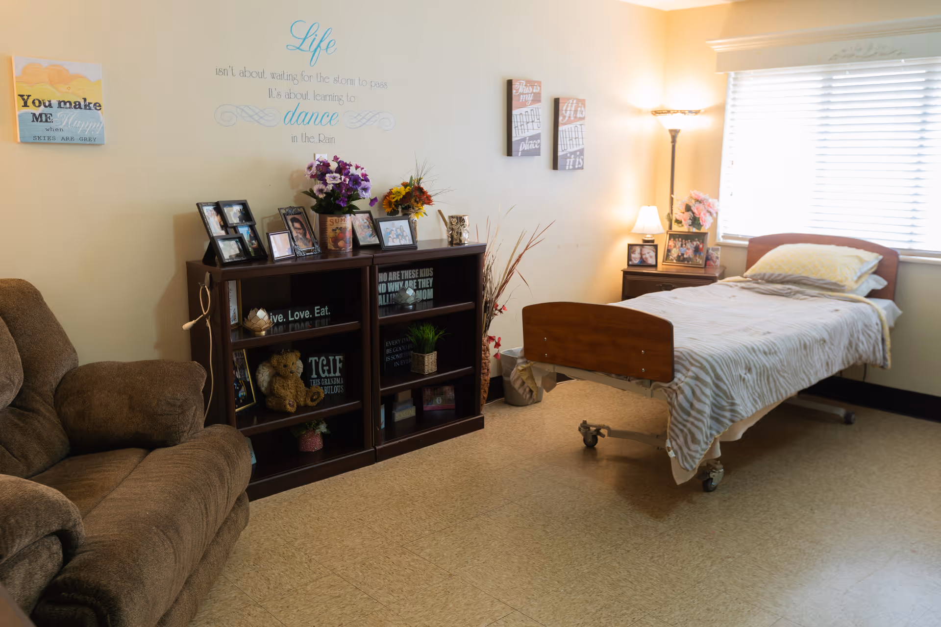 A cozy room in a senior living facility with a single bed covered in a light-colored blanket, a brown recliner chair, a dark wooden bookshelf filled with framed photos, flowers, and decorative items. The room has a window with blinds letting in natural light, a floor lamp, and a small nightstand with more framed photos and a lamp. Inspirational wall art is displayed above the bookshelf and on the walls.