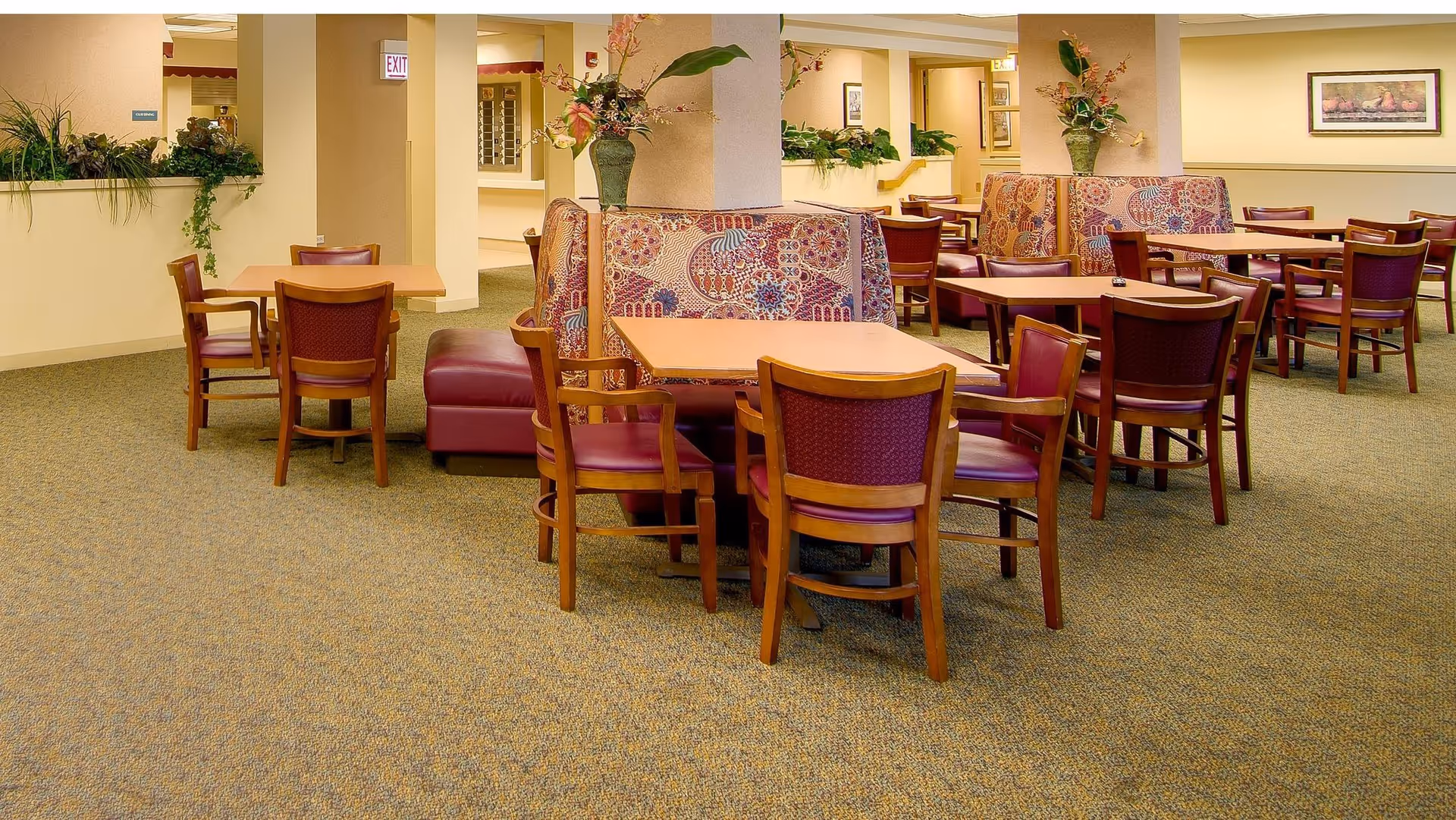 Interior view of a senior living facility dining area with multiple wooden tables and chairs arranged neatly. Some tables have patterned cushioned bench seating. The room is carpeted and decorated with potted plants and framed artwork on the walls.