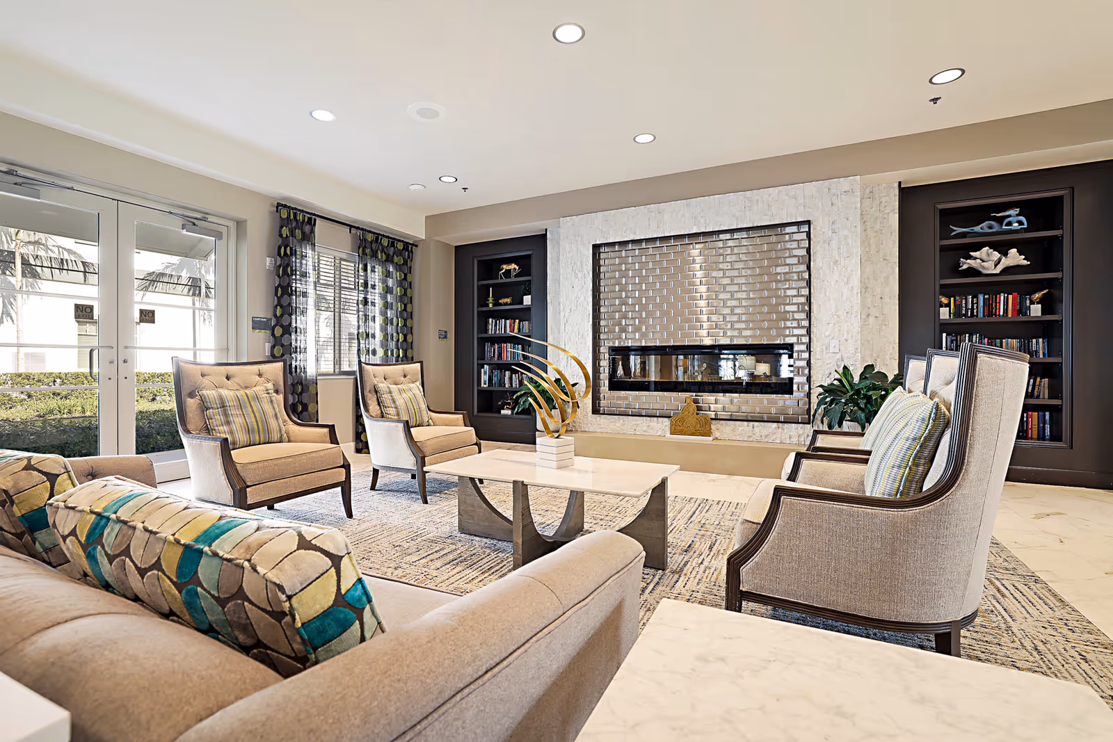 Bright furnished lounge with armchairs and a sofa arranged around a central coffee table facing a modern fireplace and built-in bookshelves.