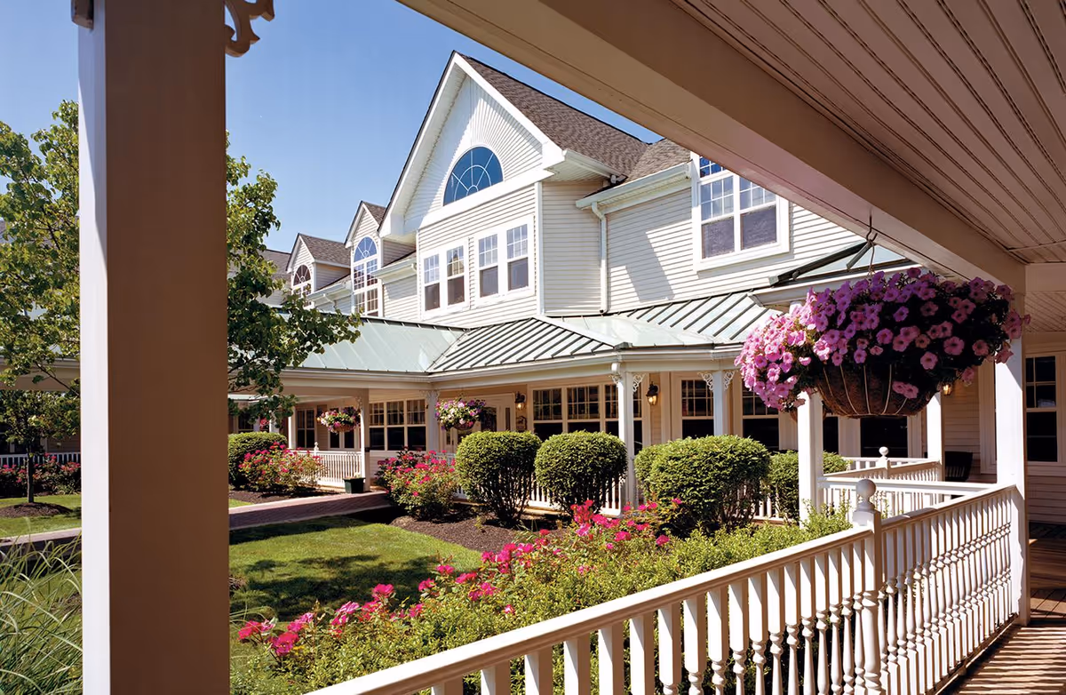 View of a senior living facility exterior with white siding, multiple windows, and a green metal roof. The foreground shows a covered porch with white railings and hanging baskets of purple flowers. There are neatly trimmed bushes and vibrant pink flowers in the landscaped garden under a clear blue sky.