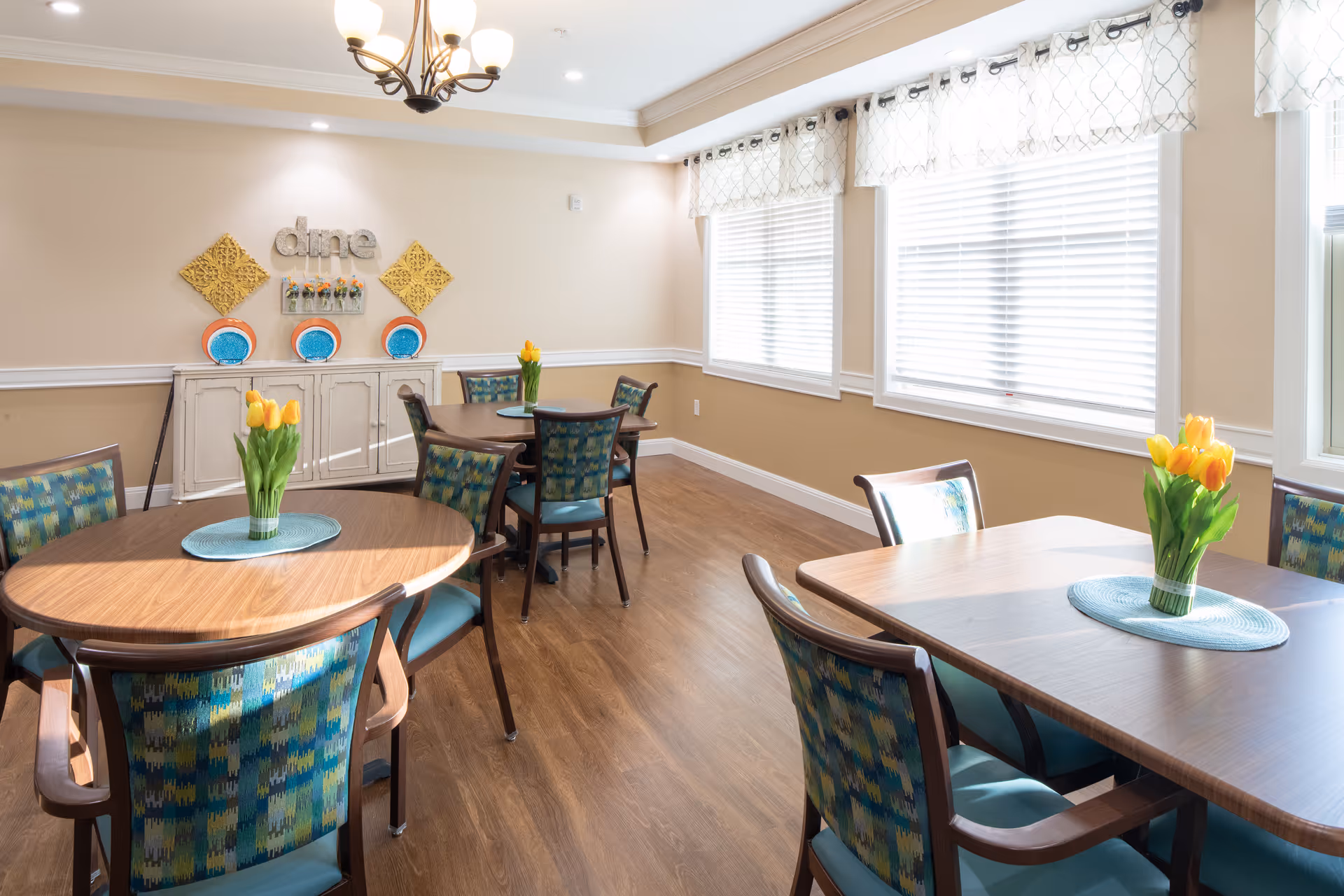 A bright dining room with wooden tables and patterned chairs. Each table has a vase with yellow tulips and a blue placemat. The room has large windows with white blinds and light-colored walls decorated with wall art and the word 'dine'.