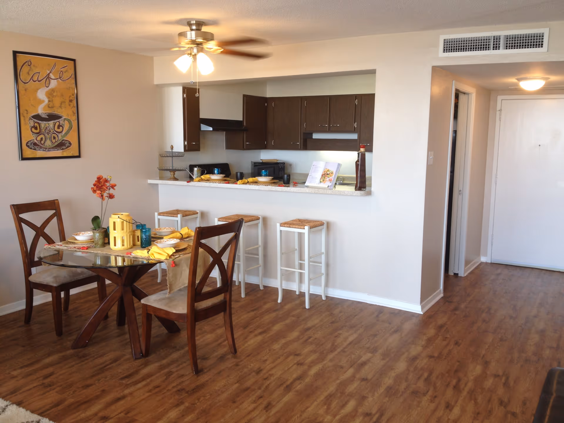 Interior view of a senior apartment dining area and kitchen. The dining area features a round glass table with four wooden chairs, set with bowls, yellow napkins, and blue glasses. A yellow decorative lantern and a small potted plant with orange flowers are on the table. The kitchen has dark brown cabinets, a countertop with four white bar stools, and various kitchen items including a cookbook and a bottle. A ceiling fan with lights is visible above, and a framed artwork with the word 'Café' and a steaming coffee cup hangs on the wall. The floor is wood-patterned, and a hallway with a white door is seen in the background.
