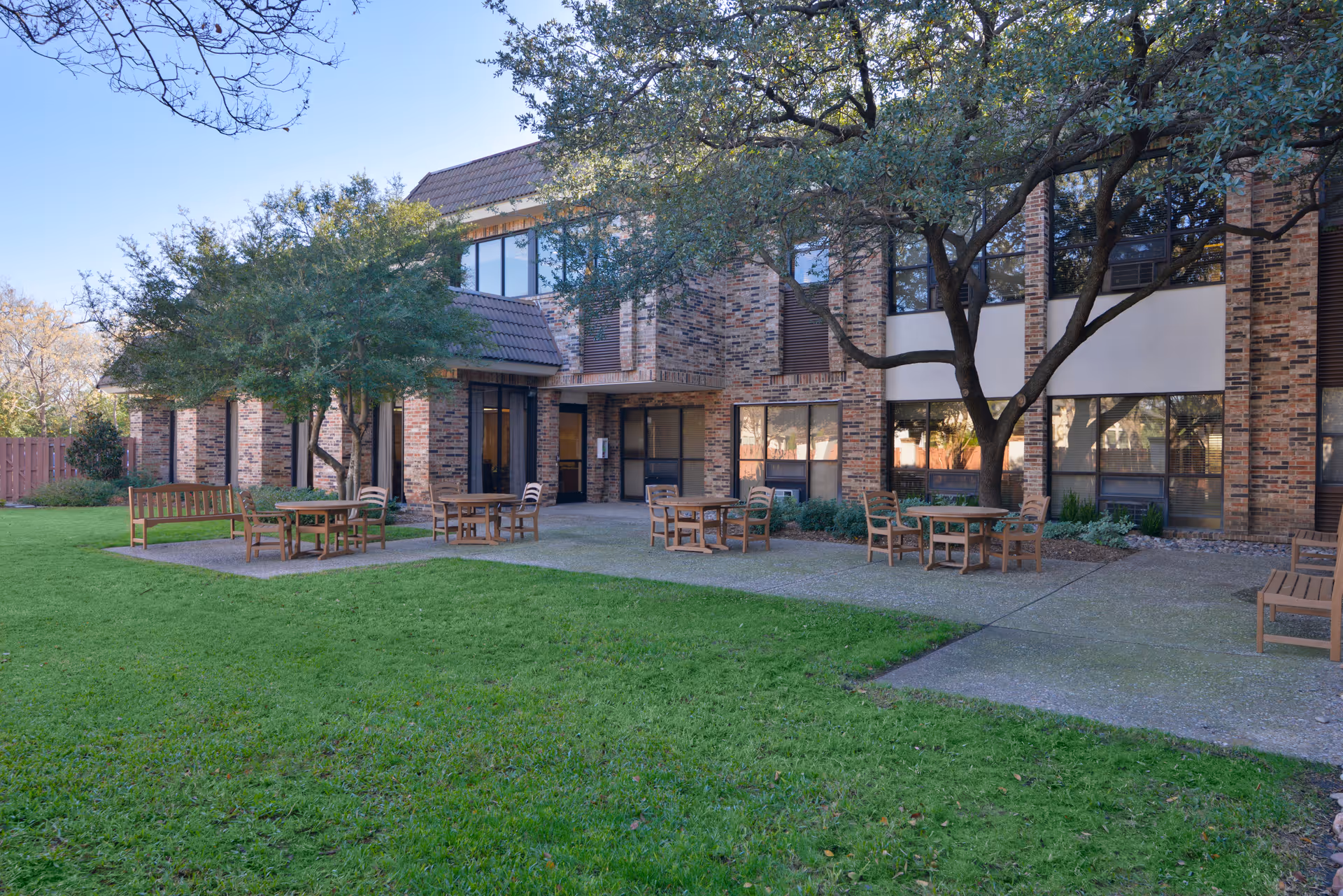 Outdoor patio area of a senior living facility with several wooden tables and chairs arranged on a concrete surface surrounded by green grass and trees. The building has a brick exterior with large windows and a tiled roof.