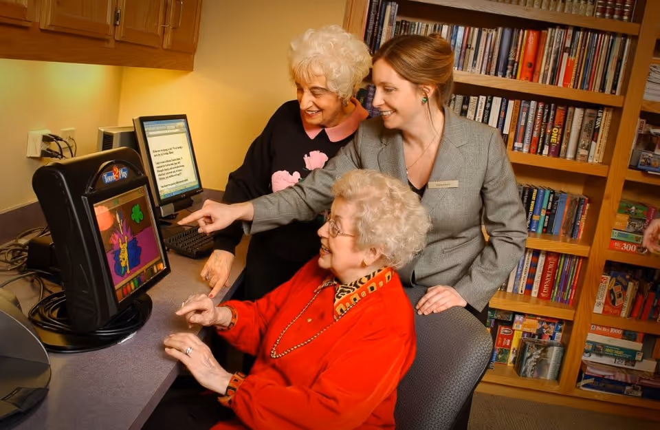 Two elderly women and a younger woman with a volunteer name tag are gathered around a computer in a room with bookshelves. One elderly woman is seated and using the computer, while the other two women are standing and pointing at the screen, smiling.