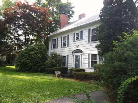 White two-story house with trimmed shrubs, trees, and a small dog on the front lawn.