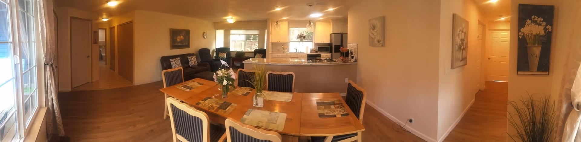 A warm, inviting interior space featuring a dining area with a wooden table and six chairs, each with dark striped cushions. The table is decorated with placemats and two vases containing flowers and greenery. Beyond the dining area is a cozy living room with a dark sofa and two armchairs, positioned near a window letting in natural light. To the right, there is a kitchen with white cabinetry, a refrigerator, microwave, stove, and a countertop with pendant lights overhead. The walls are adorned with floral artwork, and the floor is a light wood laminate. Large windows with curtains are visible on the left side, and a hallway extends from the living area toward the back of the space.