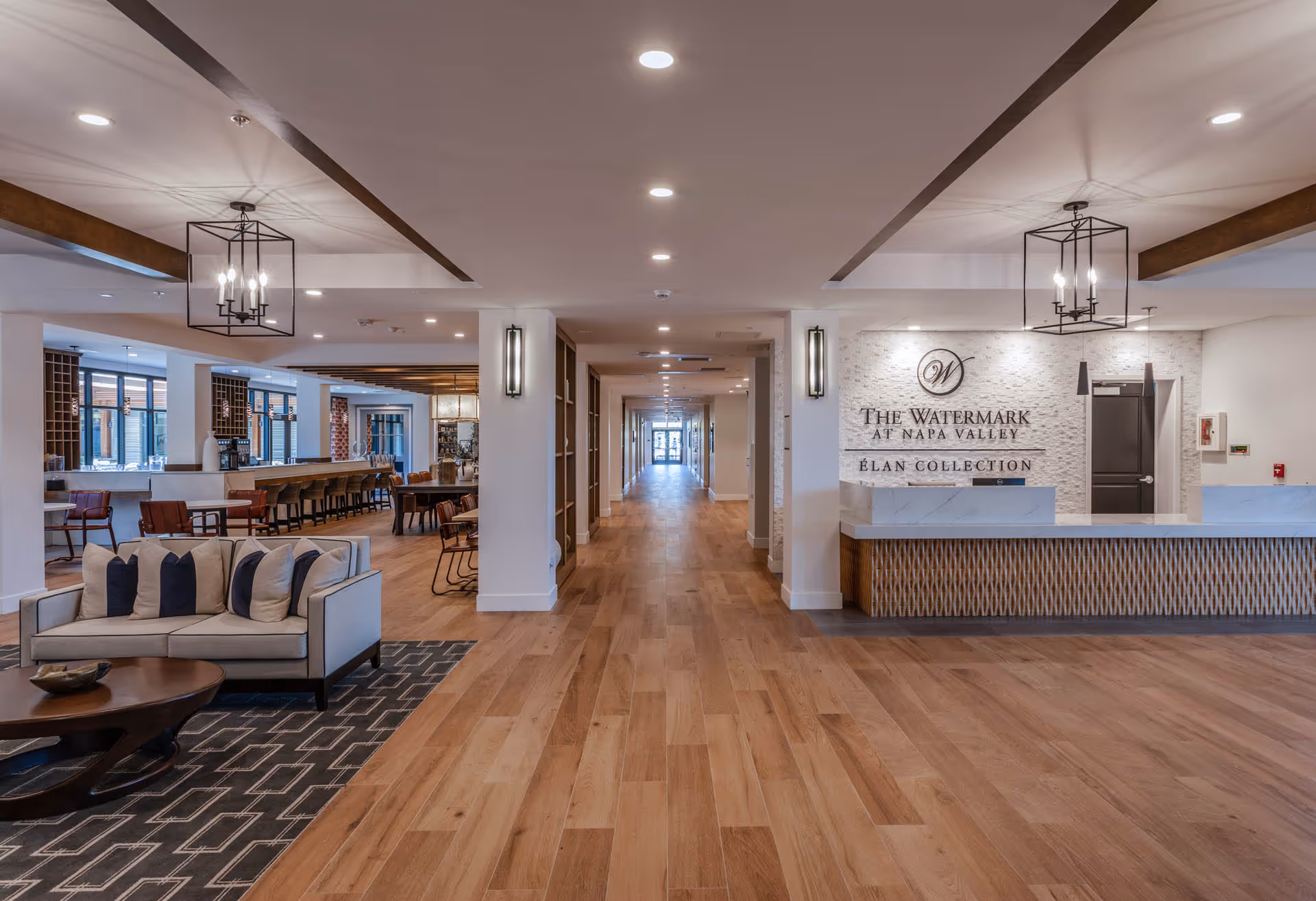 Spacious and modern interior of The Watermark at Napa Valley featuring a long wooden floor hallway, a reception desk on the right with the facility's name on the wall, a seating area with a sofa and pillows on the left, and a dining area with tables and chairs in the background.
