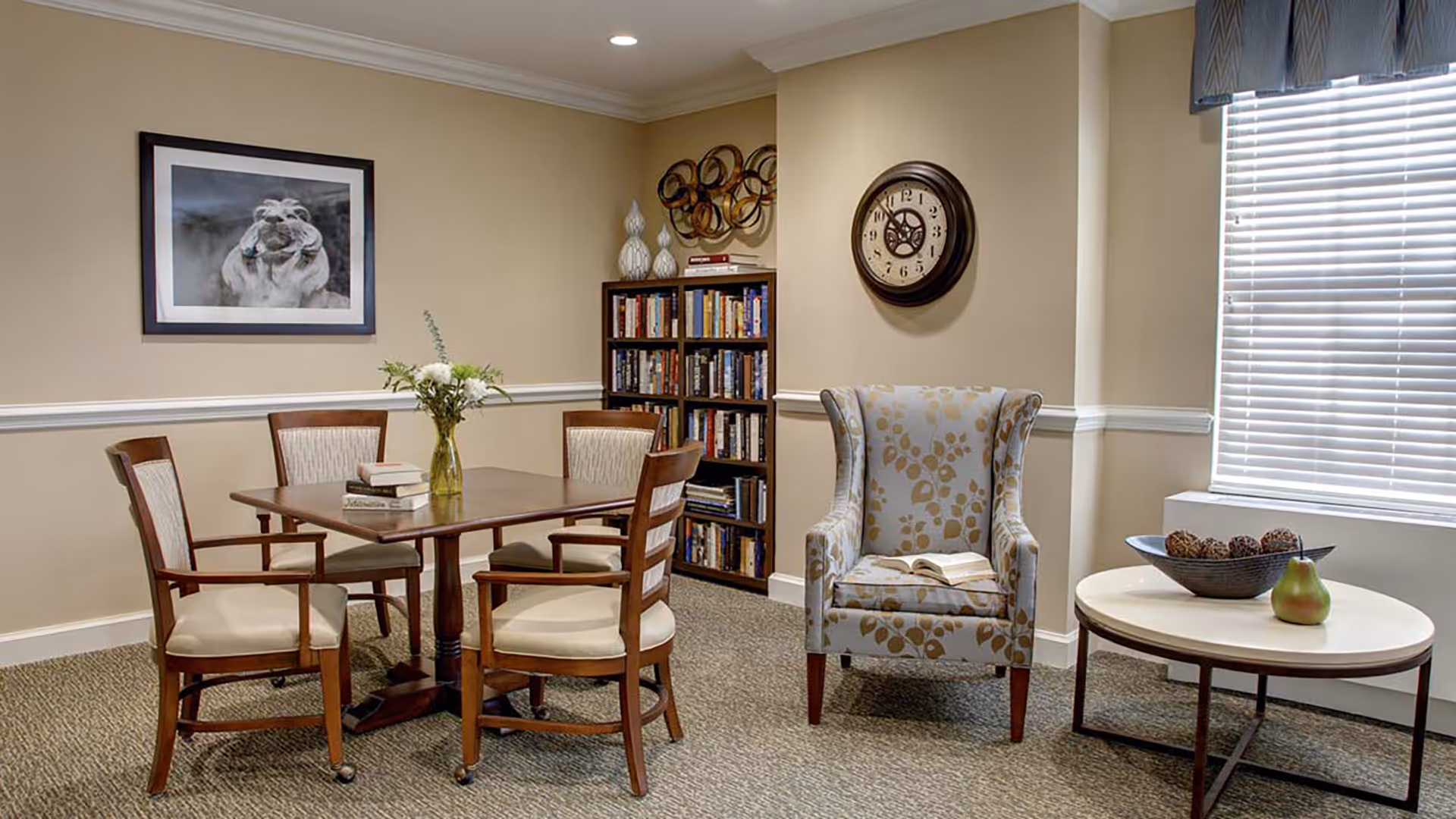 A cozy interior room with a wooden table surrounded by four cushioned chairs, a bookshelf filled with books, a patterned armchair with an open book on it, a round side table with decorative items, a large wall clock, and a framed picture of a hippopotamus on the wall.
