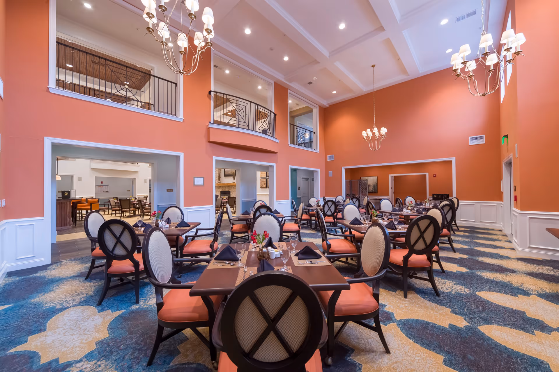 A spacious dining room with high ceilings and coral-colored walls. The room is furnished with multiple wooden dining tables, each surrounded by cushioned chairs with orange seats and beige backs. The tables are set with black napkins, glassware, and small flower arrangements. Elegant chandeliers hang from the ceiling, and there is a patterned carpet in shades of blue, beige, and orange covering the floor. Open doorways lead to adjacent rooms.