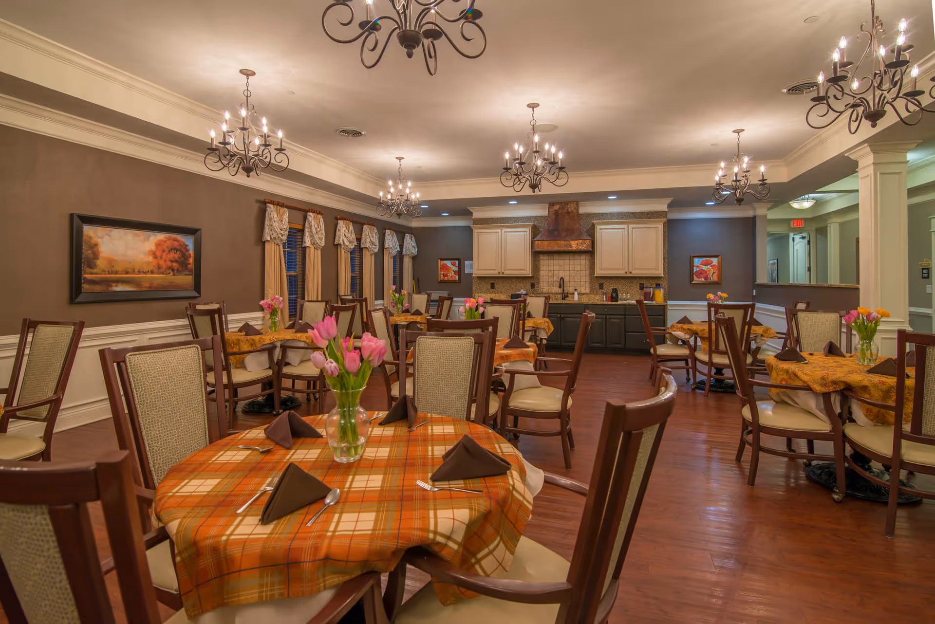 A warmly lit dining room with multiple round tables covered in orange plaid and floral tablecloths. Each table is set with brown folded napkins, silverware, and a vase of pink tulips. The room features wooden chairs with cushioned seats, chandeliers hanging from the ceiling, and a kitchen area with cabinets and a range hood in the background. The walls are decorated with paintings and windows with curtains.