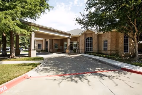 Exterior view of The Wellington at Arapaho senior living facility showing a covered entrance with brick walls, large windows, and surrounding trees casting shadows on the driveway.