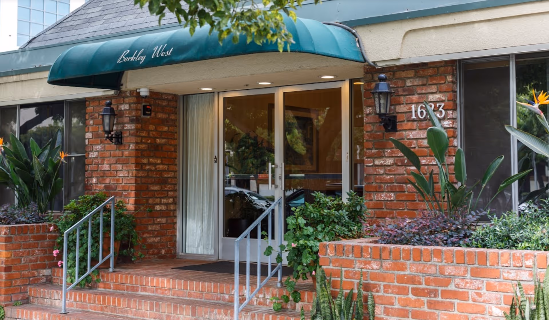 Entrance of Berkley West Healthcare Center featuring a green awning with the facility name, glass double doors, brick walls, potted plants, and a small staircase with metal railings.