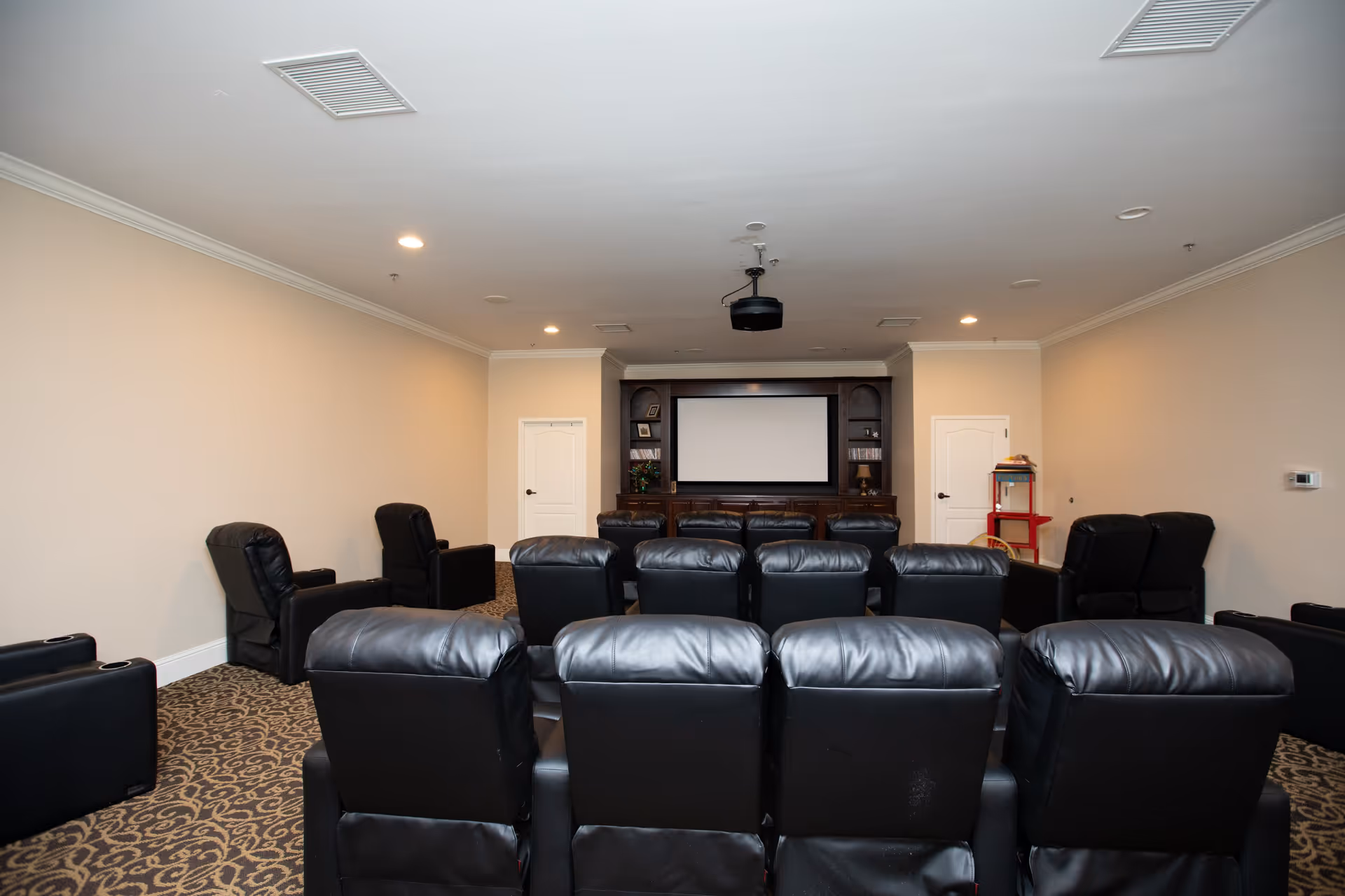 A home theater room with multiple rows of black leather recliner chairs facing a large projection screen mounted on a wooden entertainment unit. The room has beige walls, patterned carpet, recessed ceiling lights, and a popcorn machine in the back corner.