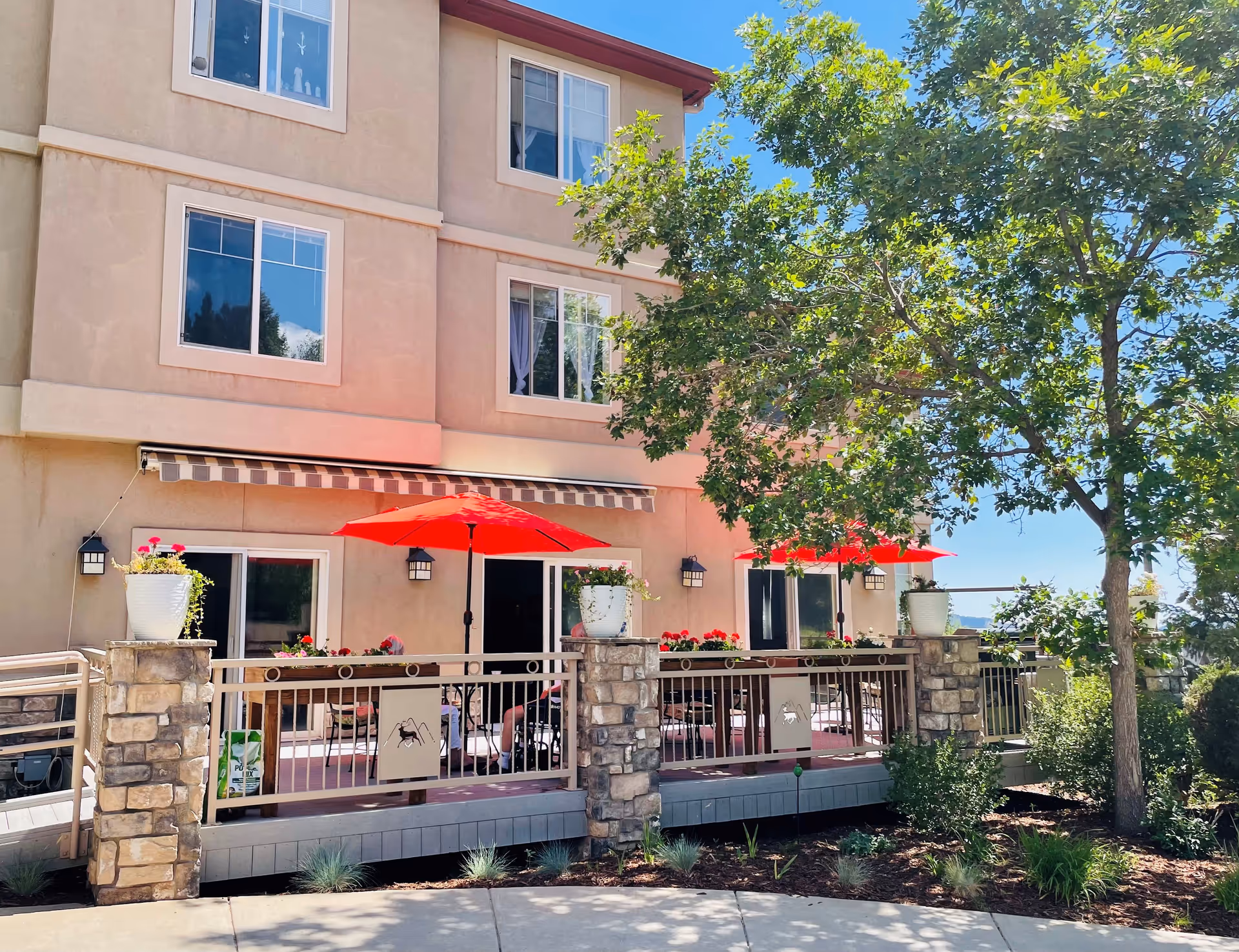 Outdoor patio area of a multi-story assisted living facility with beige walls and several windows. The patio features stone pillars, a railing, red umbrellas over tables, potted plants, and a large tree providing shade. The sky is clear and blue.