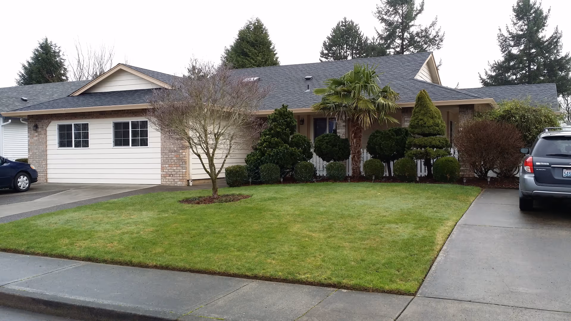Front exterior view of a single-story residential house with a well-maintained lawn, various shrubs, a small tree, and two parked cars on the driveway.