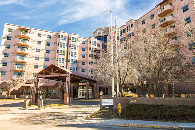 Exterior view of a multi-story senior living facility building with a covered entrance, leafless trees, and a sign that reads Brookdale University Park. The sky is partly cloudy.