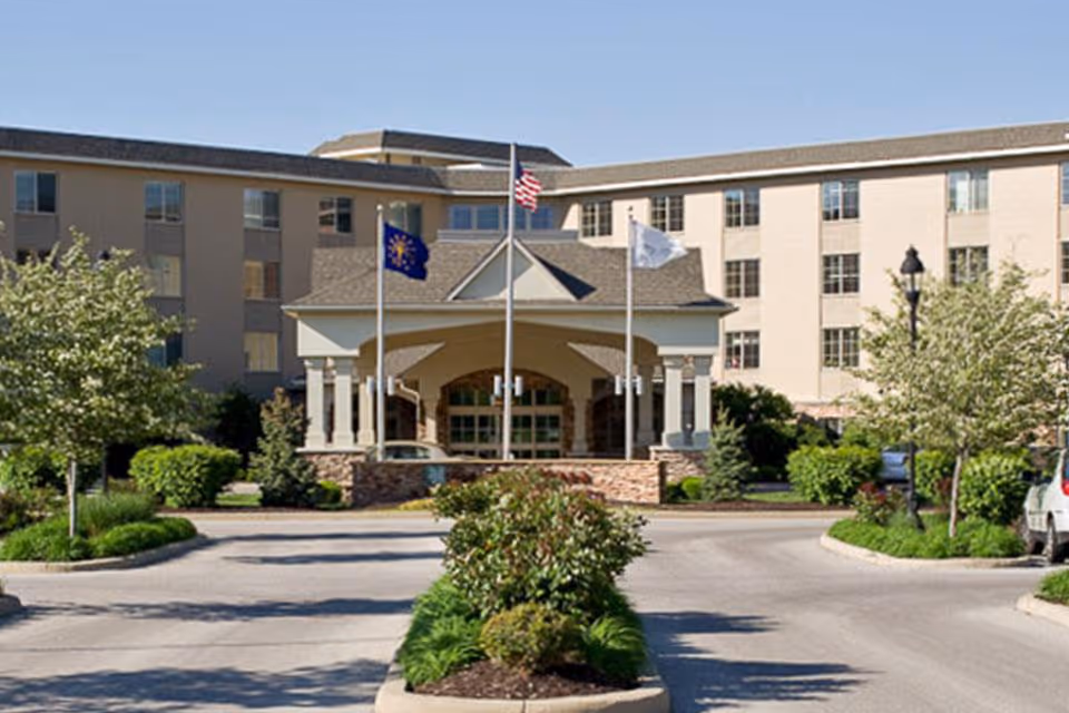 Front exterior view of Westside Village Nursing Center showing a multi-story building with a covered entrance, three flagpoles with flags, landscaped greenery, and a circular driveway.