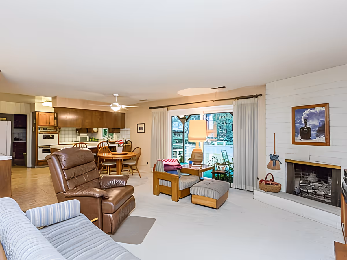 Open living room with a leather recliner, sofa, ottoman and fireplace, flowing into a dining area and kitchen with sliding glass doors to a patio.
