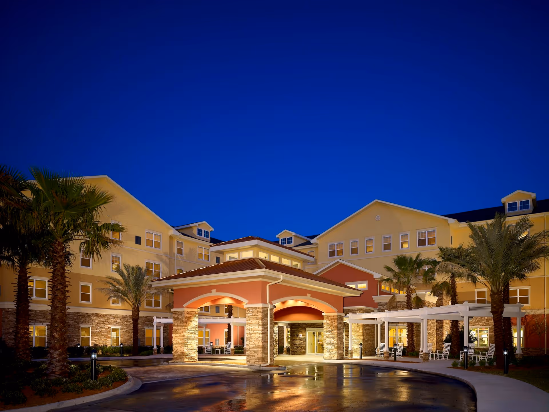 Exterior view of Camellia At Deerwood senior living facility at dusk, showing a large building with multiple windows, palm trees, and a covered entrance with stone pillars and warm lighting.