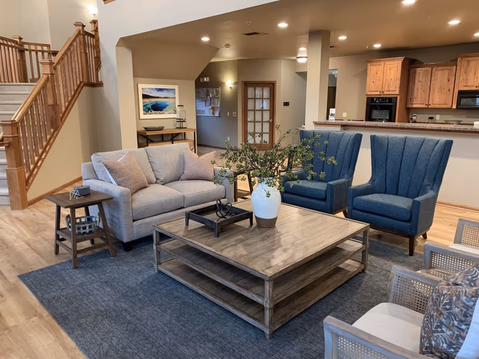 A cozy living room area in Autumn Springs Assisted Living featuring a light gray sofa with two pillows, two blue upholstered armchairs, and two wooden chairs with cushions. A large wooden coffee table with a white vase holding green branches sits in the center on a blue rug. In the background, there is a kitchen with wooden cabinets and a staircase with wooden railings leading upstairs.