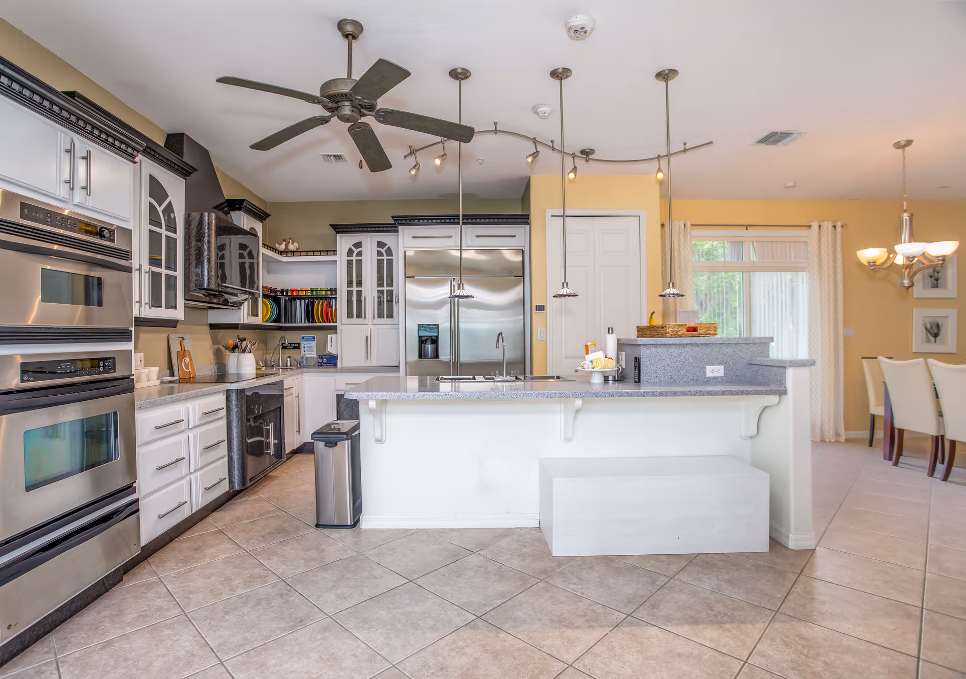 Modern kitchen with white cabinetry, stainless steel double ovens, a large stainless steel refrigerator, a kitchen island with a sink, pendant lights hanging from the ceiling, a ceiling fan, and a dining area visible in the background with a chandelier and white chairs.