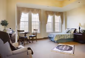 Bright furnished senior living bedroom with a bed, nightstands, an armchair, a small table and chairs, and large draped windows.
