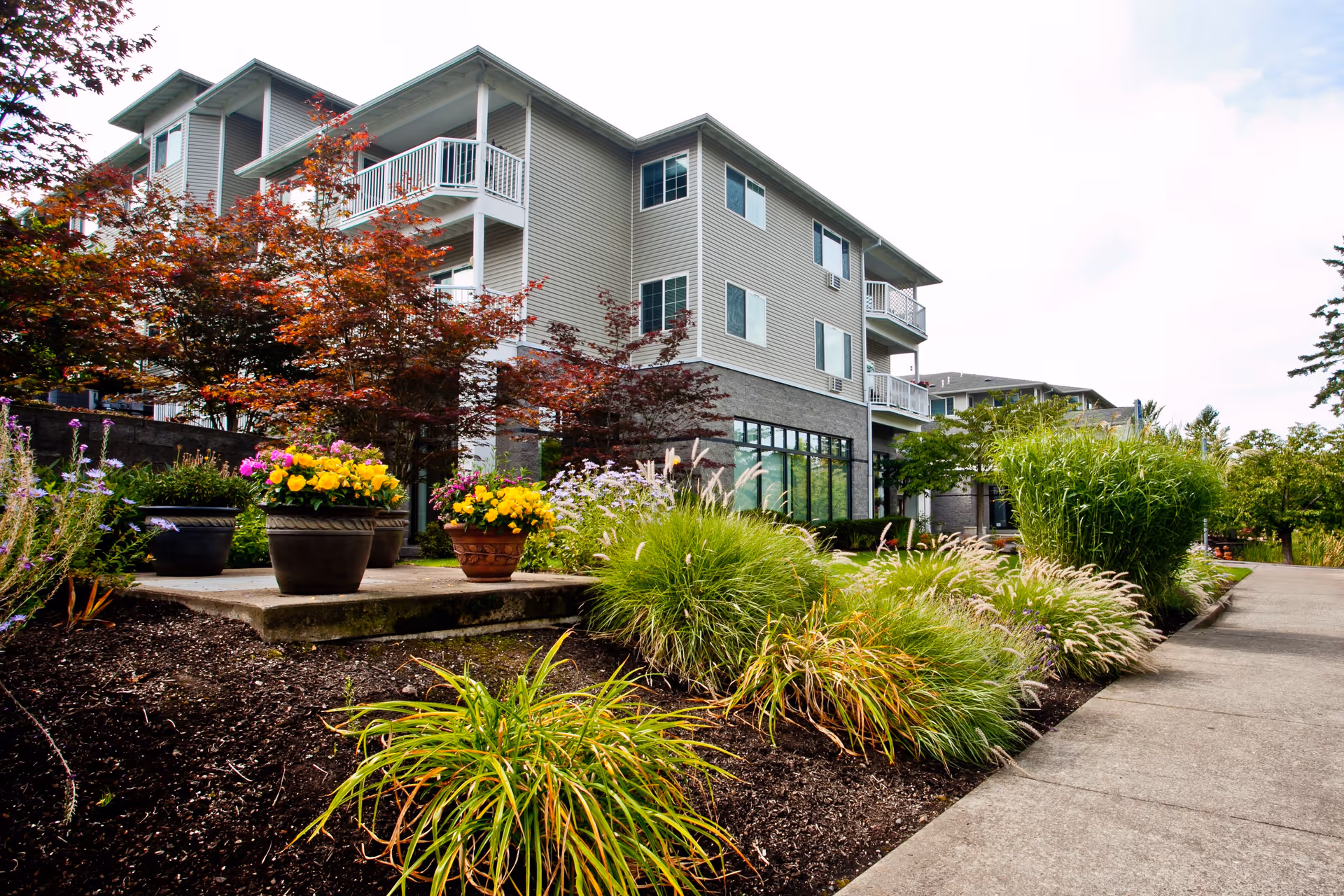 Exterior view of a multi-story residential building with balconies, surrounded by landscaped gardens featuring colorful flowers, shrubs, and ornamental grasses along a concrete walkway.