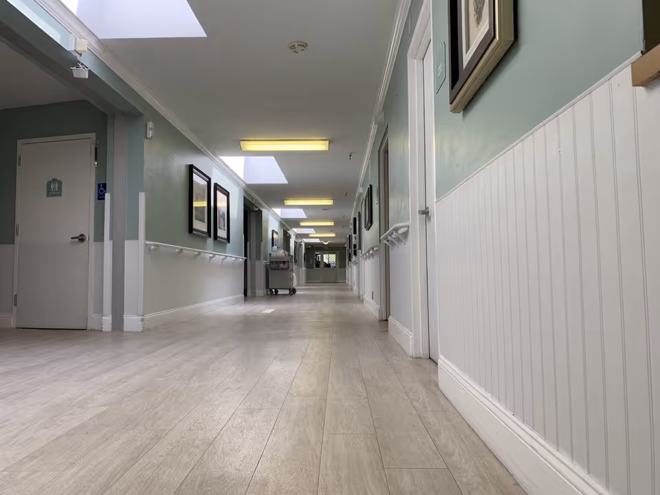 Long bright interior hallway in a senior living facility with handrails, skylights, framed pictures, and light wood flooring.