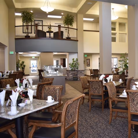 A spacious dining room with multiple tables set with white napkins, cups, and small flower arrangements. The room features high ceilings, large columns, and a mezzanine level with plants. The decor includes patterned chairs and carpeted floors, with natural light coming through windows and glass doors in the background.