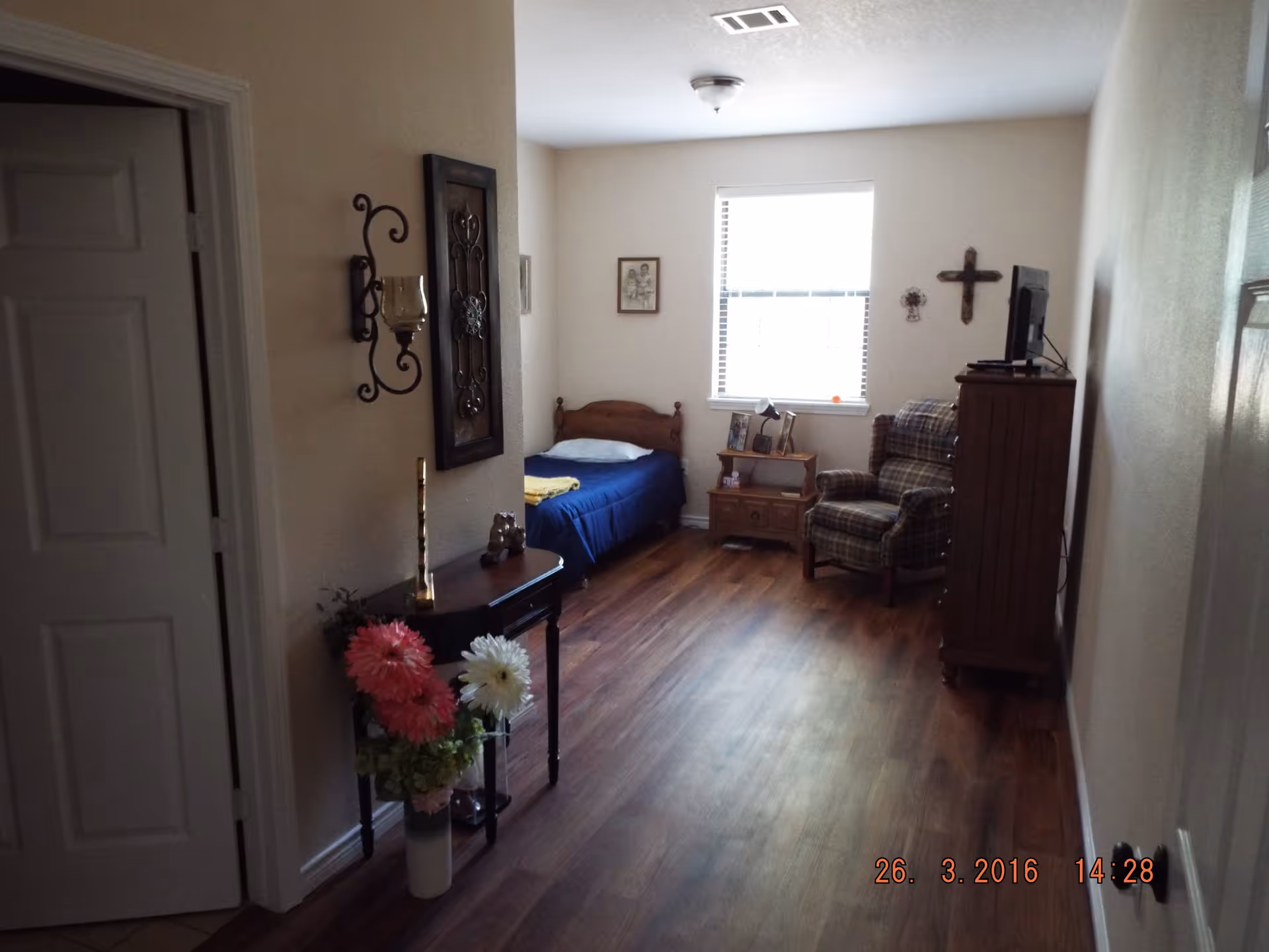 A small bedroom with a single wooden bed covered with a blue blanket and a yellow towel on top. There is a wooden nightstand with framed photos and a lamp next to the bed. A plaid upholstered armchair is positioned near the window, which has white blinds. On the wall above the armchair is a wooden cross and a small decorative item. The floor is wooden, and there is a small table with flowers and decorative items near the entrance. The walls are painted light beige.