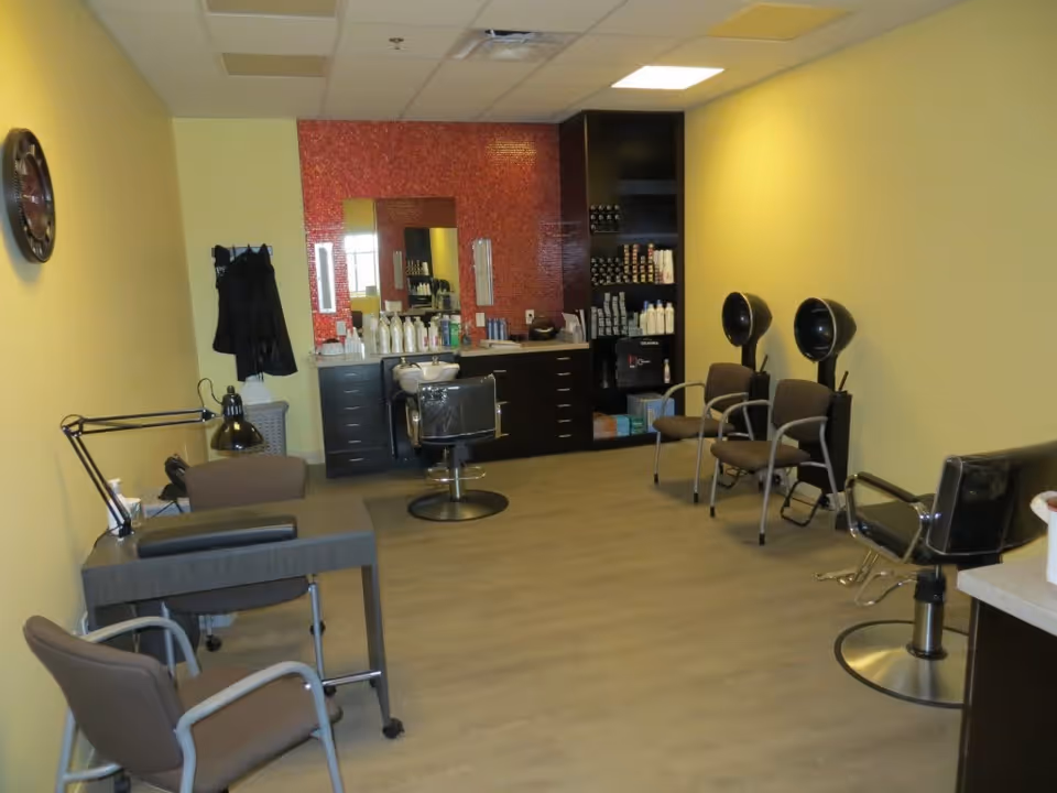 Interior of a salon room with yellow walls and wood flooring. The room contains salon chairs, hair drying stations, a manicure table with a lamp, and shelves stocked with hair care products. A large mirror is mounted on a red tiled wall behind the main salon chair.