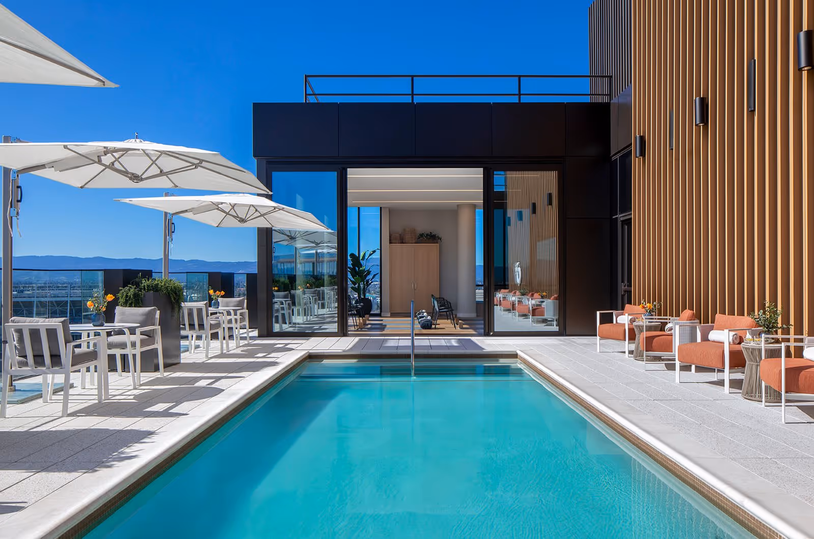 Outdoor rooftop pool area with clear blue water, surrounded by white lounge chairs and tables with white umbrellas on the left side, and orange cushioned chairs with small tables on the right side. The background shows a modern building facade with vertical wooden slats and large glass doors leading to an indoor space with more seating and plants. The sky is clear and blue with distant mountains visible.