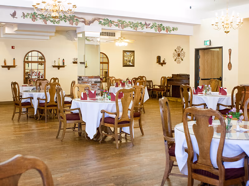 Spacious dining room with round tables set with white tablecloths, wooden chairs, and chandeliers.