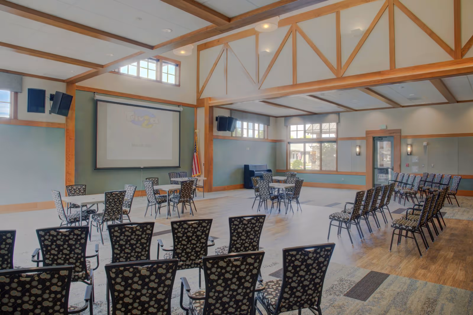Spacious multipurpose common room with rows of patterned chairs, round tables, a projector screen, high beamed ceiling and large windows.