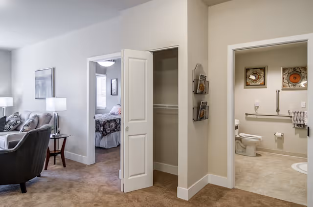 Interior view of a senior living facility showing a living room area with a gray armchair, a side table with a lamp, and a sofa with cushions. There is an open doorway leading to a bedroom with a bed covered in a floral bedspread. Another open doorway reveals a bathroom with a toilet, grab bars, and framed artwork on the wall. A closet with a bi-fold door is also visible in the center.