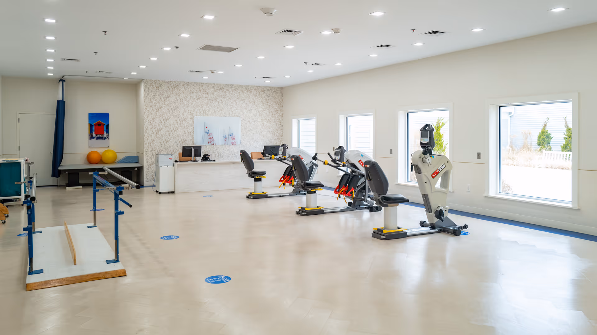 A spacious rehabilitation or physical therapy room with exercise equipment including stationary bikes and parallel bars. The room has large windows letting in natural light, a desk with computers, and a clean, bright interior.