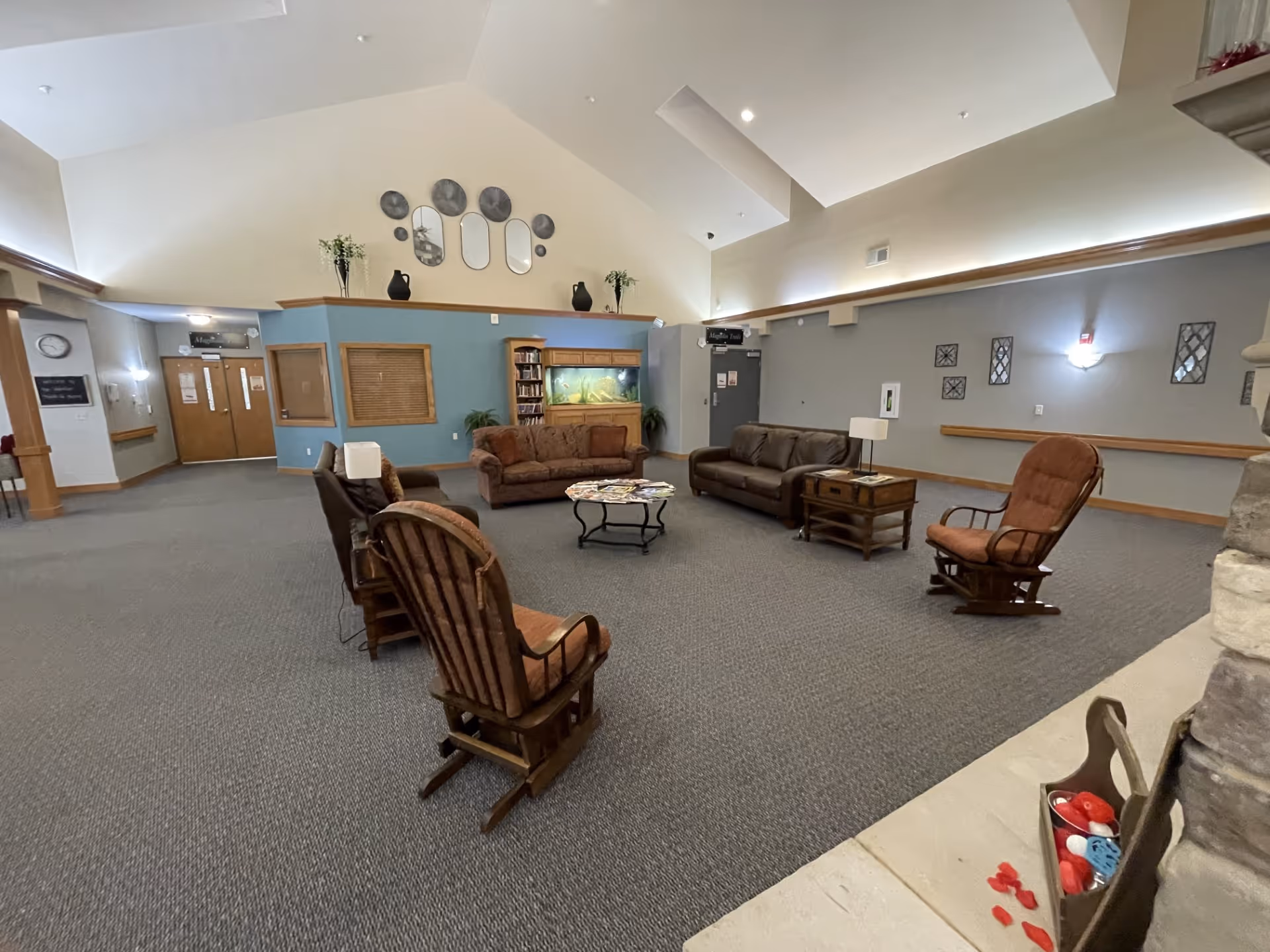A spacious senior living facility common area with high vaulted ceilings, carpeted floor, and neutral-colored walls. The room features several cushioned sofas and wooden rocking chairs arranged around a central coffee table with magazines. There is a large fish tank against a blue accent wall, decorative mirrors and vases on a ledge above, and wall-mounted lights providing soft illumination. A stone fireplace is partially visible on the right side.