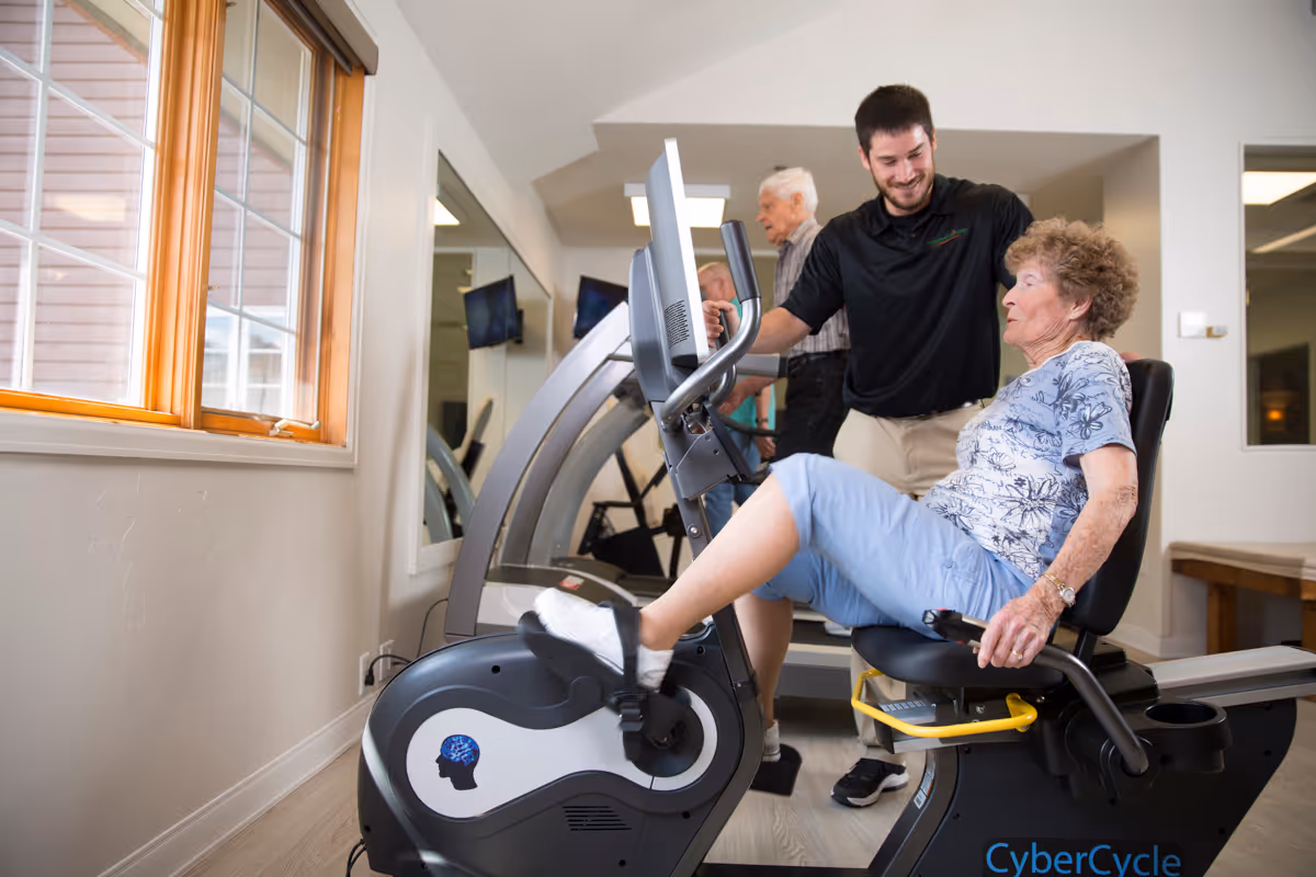 An elderly woman exercises on a recumbent stationary bike inside a fitness room, assisted by a smiling male staff member. In the background, two other elderly individuals use treadmills. The room has large windows and mirrors on the walls.