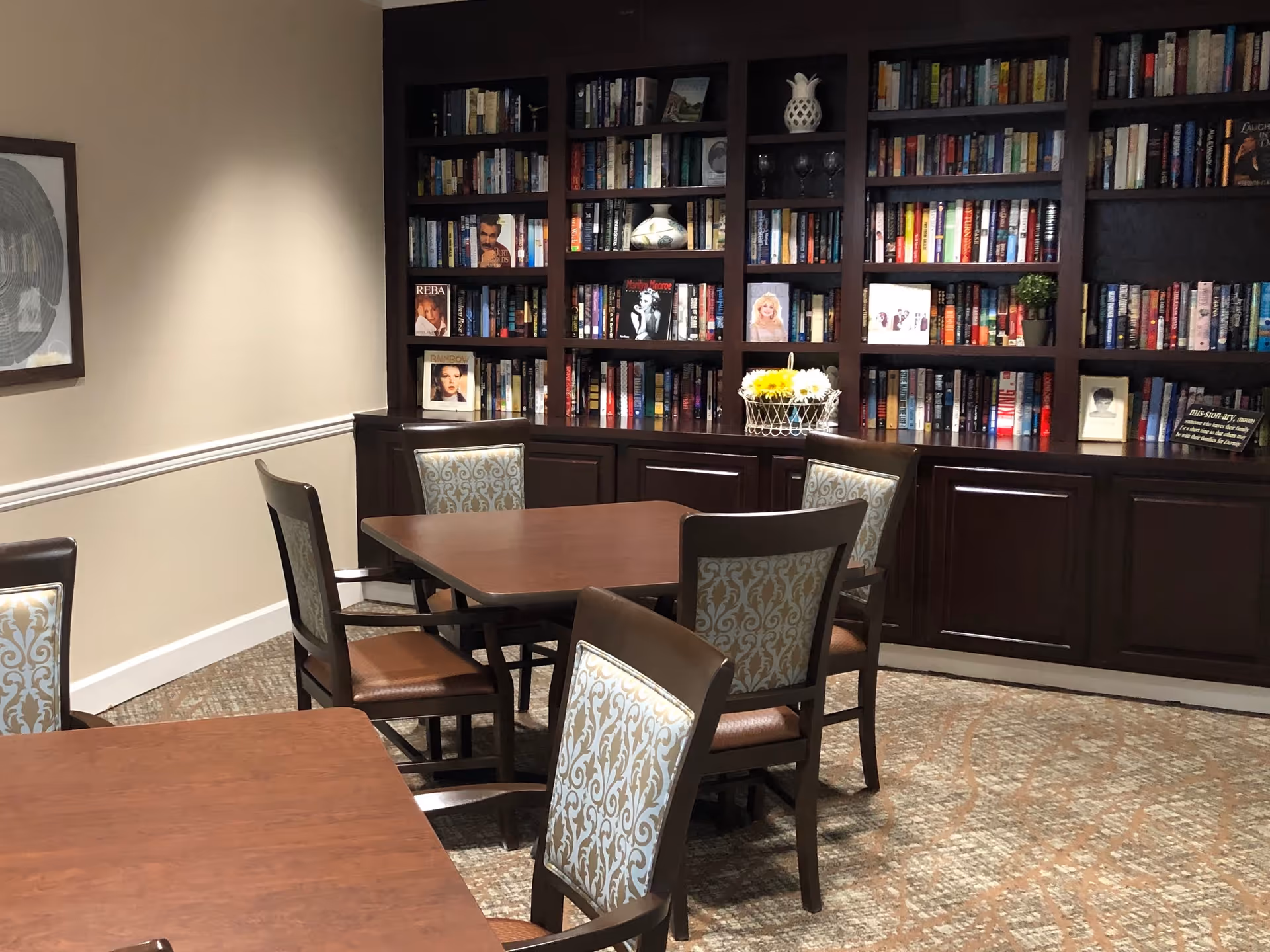 Interior room with wooden tables and chairs featuring patterned upholstery. A large dark wood bookshelf filled with books and decorative items covers the back wall. The room has beige walls with white trim and a carpeted floor.