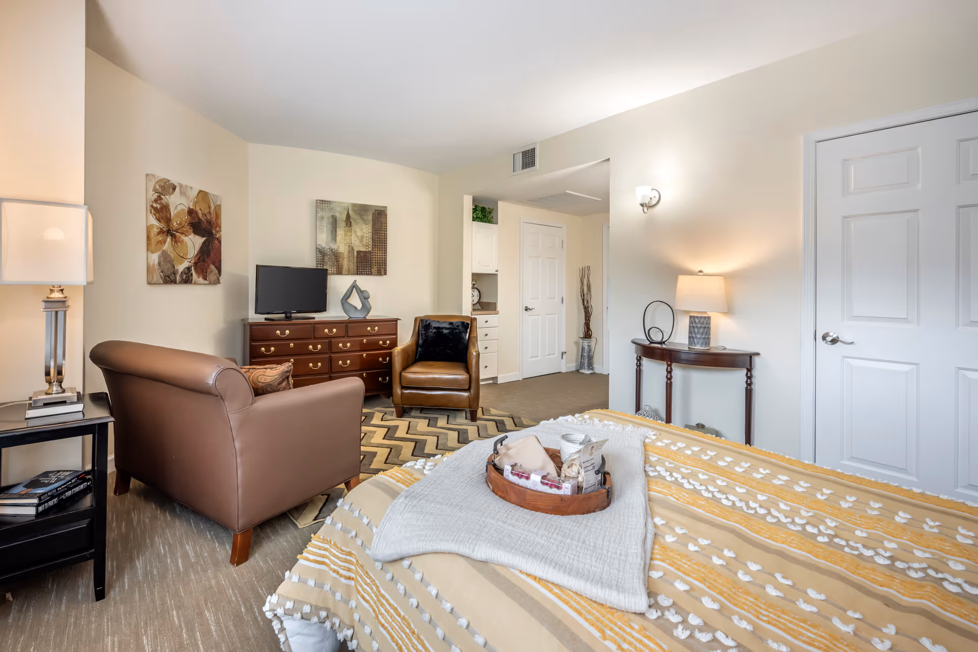 A cozy bedroom in a senior living facility with a bed covered in a yellow and white patterned blanket. A tray with cups and snacks is placed on the bed. The room features two brown leather chairs, a wooden dresser with a TV on top, a side table with a lamp, and wall art. The walls are painted light beige, and there is a door and a small hallway visible in the background.