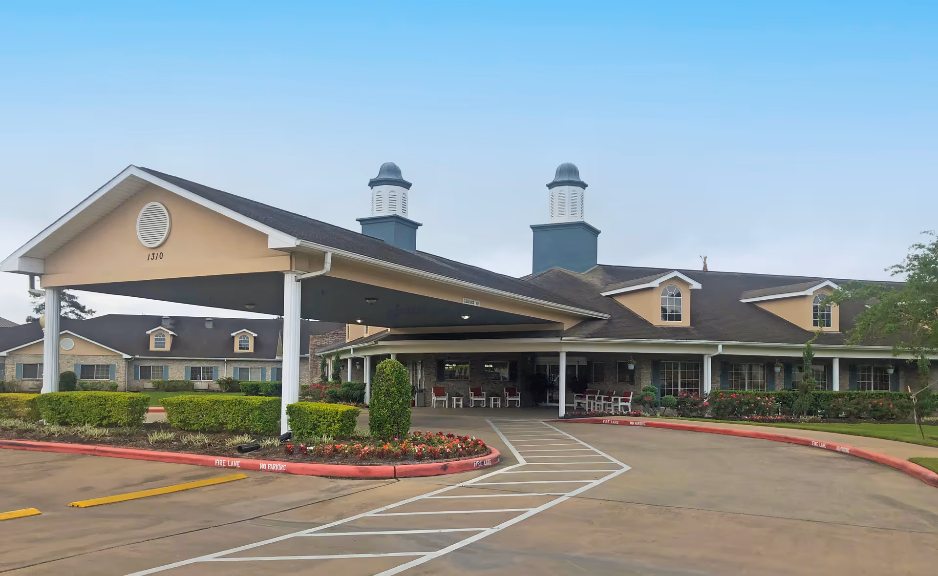 Front entrance of a single-story senior living facility with a covered porte-cochere, landscaped flower beds, and a wraparound porch with chairs.