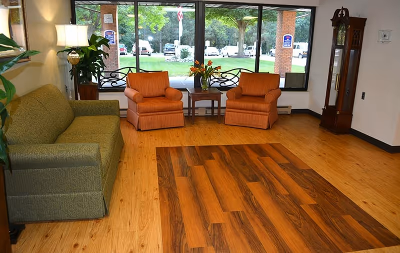 Seating area with a green sofa, two orange armchairs and a small table in front of large windows overlooking the facility grounds.