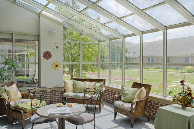 A bright sunroom featuring comfortable seating, plants, and large windows overlooking a garden area.