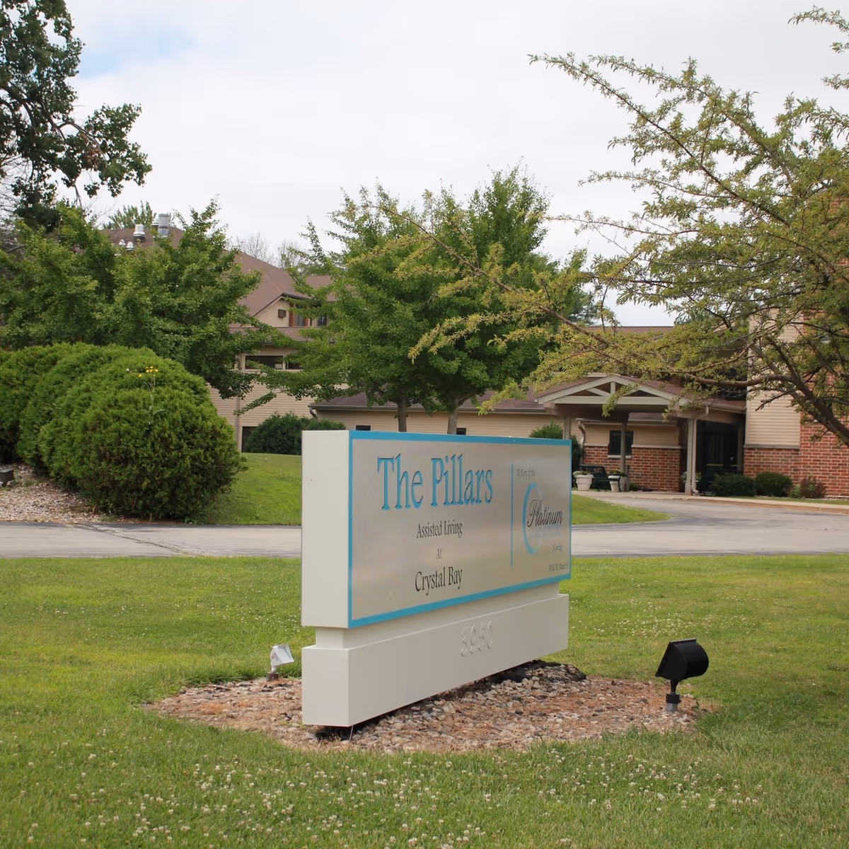A lawn sign reading "The Pillars" in front of a senior living building with trees, shrubs, and a driveway.