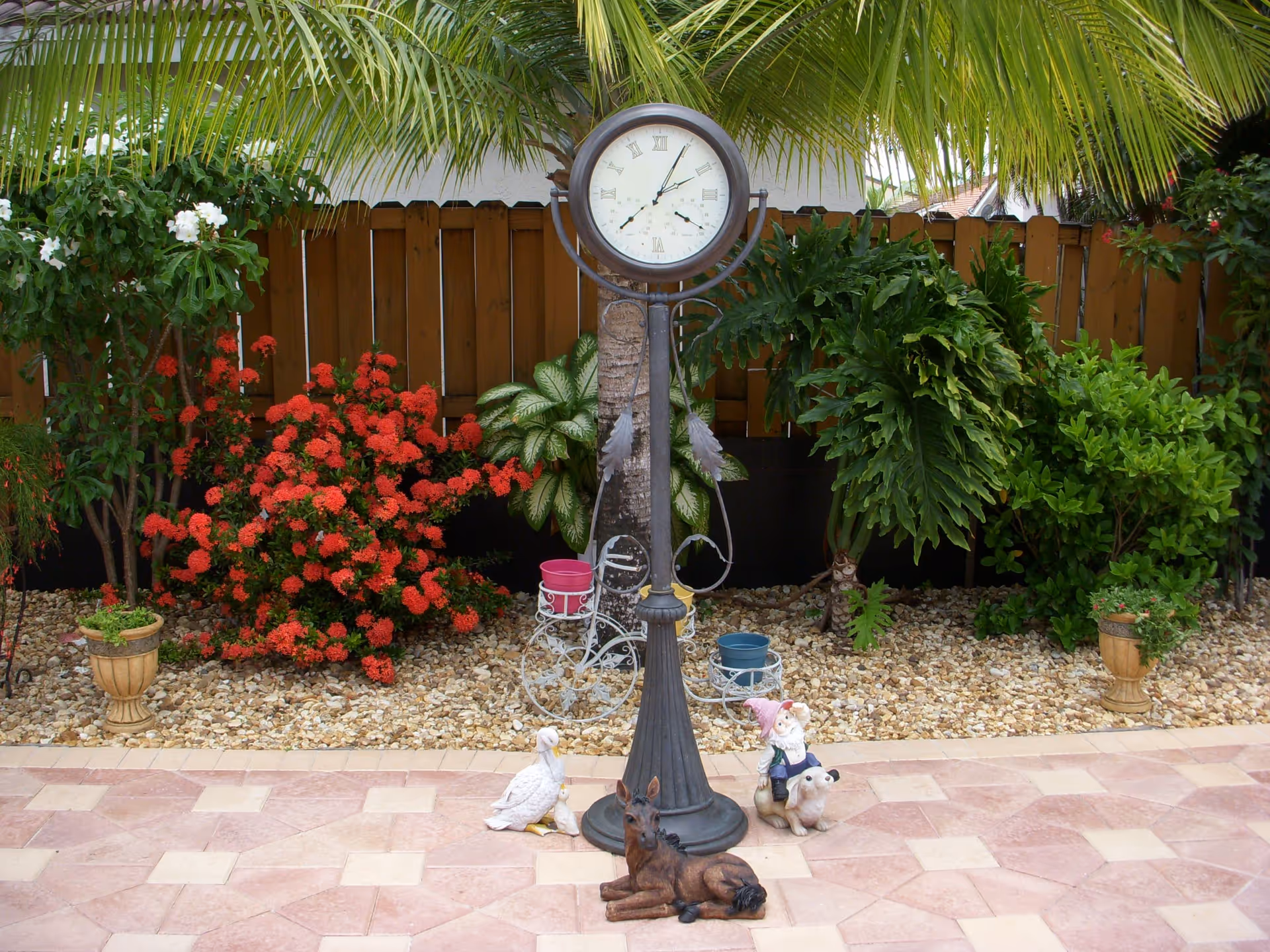 Outdoor garden area with a tall standing clock in the center surrounded by decorative plants and garden ornaments including a duck, a dog, and a gnome. There are various green plants and red flowers along a wooden fence in the background, with a tiled patio in the foreground.
