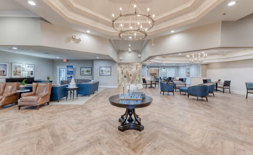 Spacious and well-lit senior living facility common area with a round wooden table in the center decorated with a welcome sign, vases, and candles. Surrounding the table are multiple seating areas including brown leather armchairs, blue sofas, and several tables with chairs. The room features modern chandeliers, light wood flooring, and neutral-colored walls adorned with framed artwork.