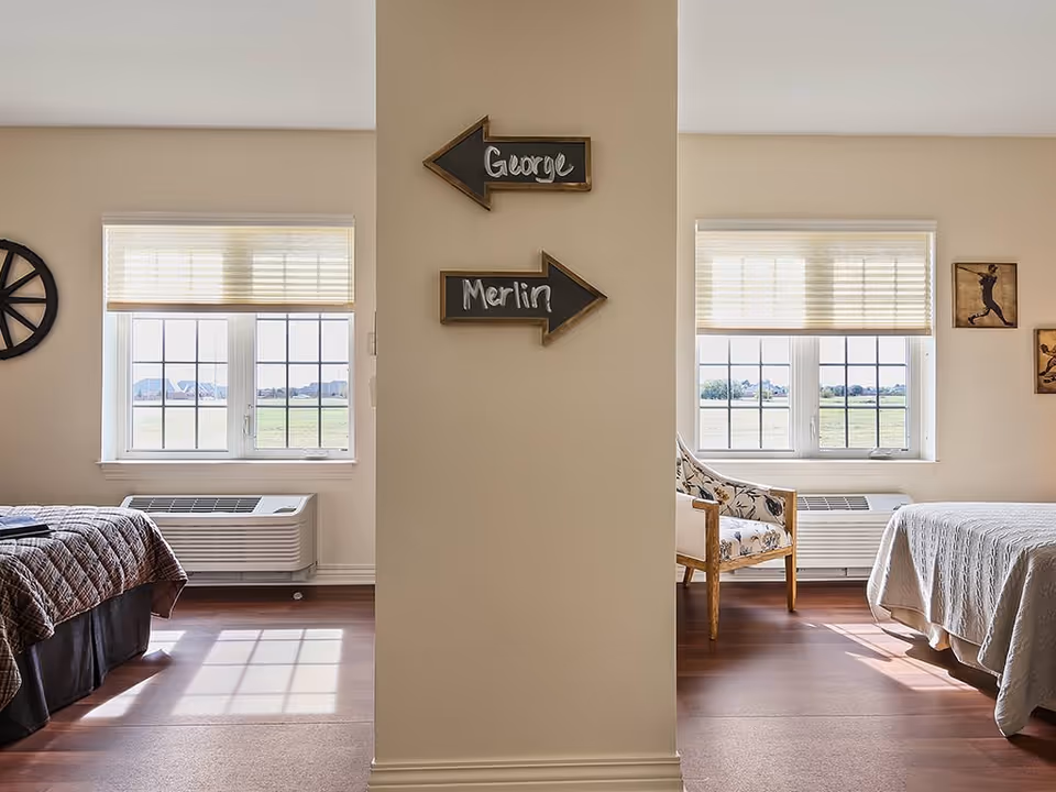 Interior view of a senior living facility room divided by a partial wall with two arrow signs labeled 'George' pointing left and 'Merlin' pointing right. Each side has a bed, a window with blinds, and an air conditioning unit below the window. The left side has a dark quilted bedspread and a decorative wheel on the wall, while the right side has a light bedspread, a floral armchair, and framed artwork on the wall.