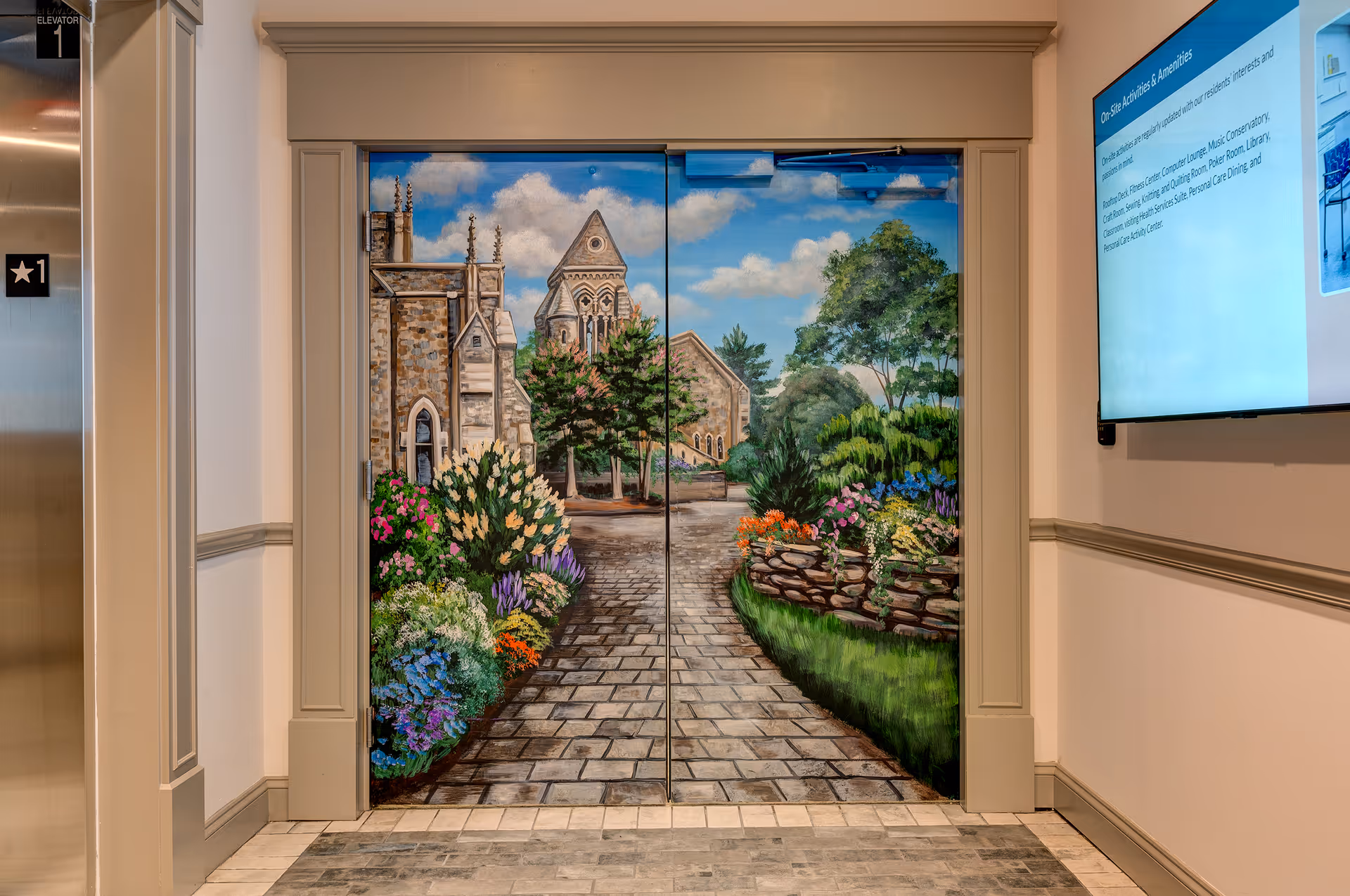 Elevator doors painted with a mural depicting a stone pathway leading to a historic stone building surrounded by colorful flowers and greenery under a blue sky with clouds. To the left of the doors is a stainless steel elevator panel with floor indicators, and to the right is a wall-mounted digital screen displaying information about on-site activities and amenities.