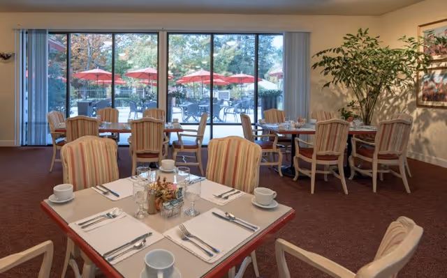 Dining room with several tables set for four people each, featuring striped cushioned chairs, white cups, glasses, and silverware. Large sliding glass doors in the background open to an outdoor patio area with tables and red umbrellas. A large potted plant and framed artwork decorate the interior space.