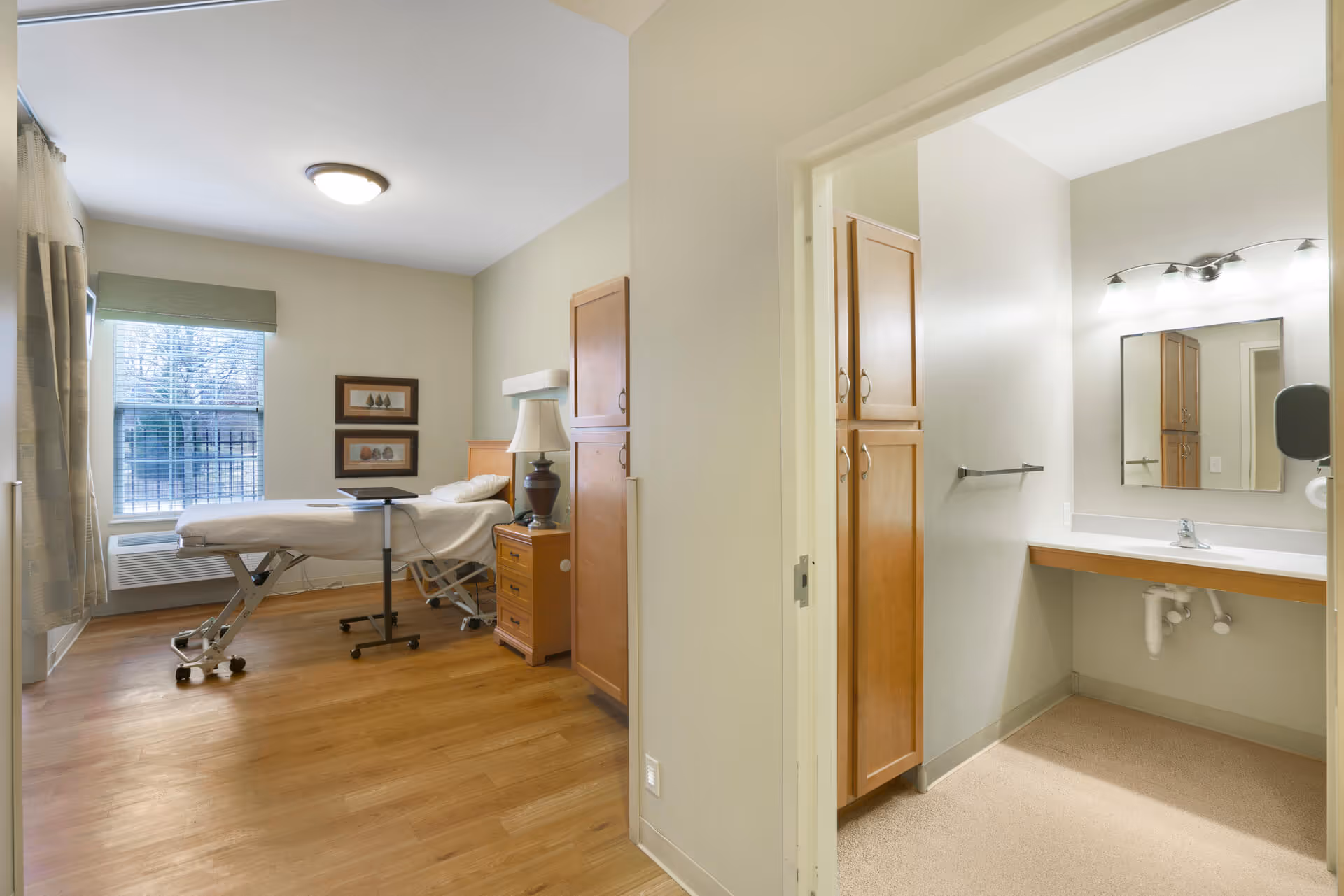 A senior living facility room with a hospital-style bed on wheels, a bedside table with a lamp, and two framed pictures on the wall. The room has a large window with blinds and curtains, and wooden flooring. Adjacent to the room is a small bathroom area with a sink, mirror, wooden cabinets, and a towel rack.